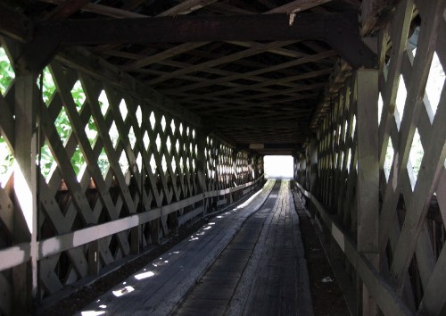 A view inside a covered bridge, featuring wooden beams and a light-filled exit at the far end.
