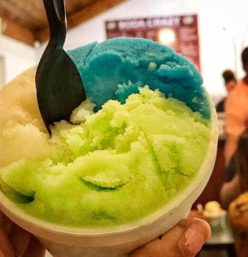 A hand holding a cup of colorful sorbet with blue, green, and white layers, featuring a black spoon.