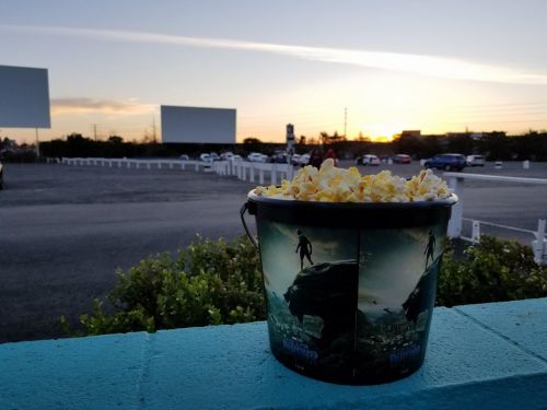 A bucket of popcorn sits on a ledge with a sunset and drive-in movie screens in the background.