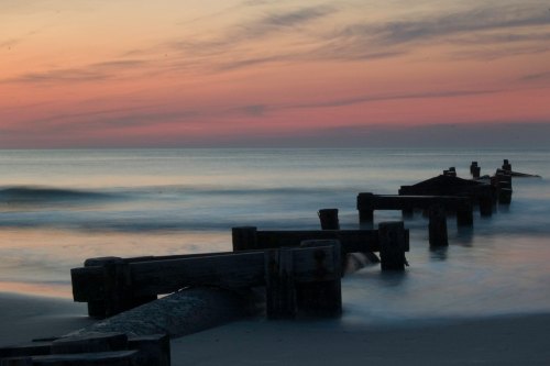 A serene beach scene at sunset, featuring weathered wooden posts extending into calm waters under a colorful sky.