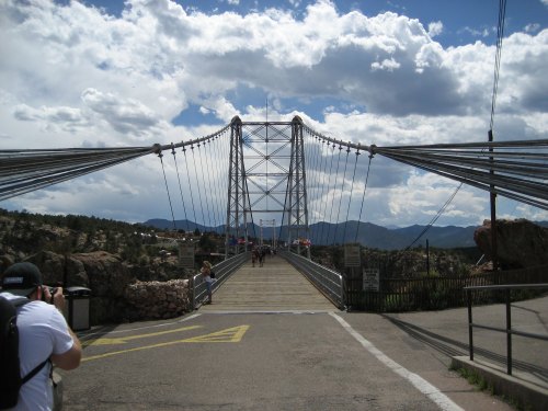 A suspension bridge spans a rocky landscape under a cloudy sky, with people walking across and mountains in the background.