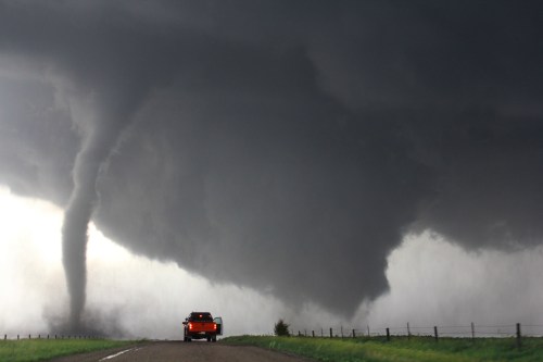 A tornado forms in a stormy sky, with a truck parked on a road nearby, surrounded by green grass and a fence.