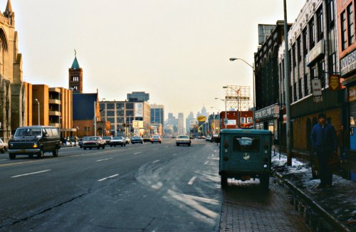 A city street scene with vintage cars, buildings, and a distant skyline under a gray sky. Snow is visible on the ground.