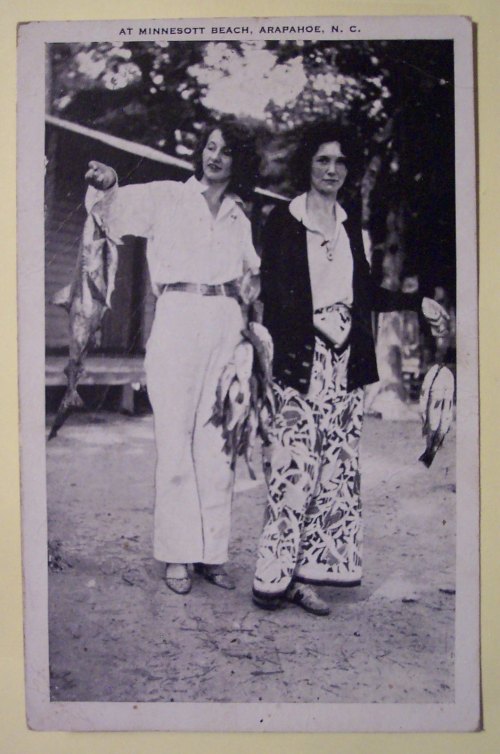 Two women holding fish at Minnesott Beach, Arapahoe, N.C., dressed in vintage clothing, smiling outdoors.