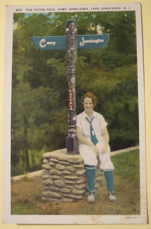 A woman in white attire sits beside a colorful totem pole marked "Camp Junaluska" in Lake Junaluska, NC.