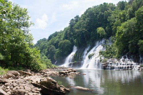 A serene waterfall cascades down rocky cliffs, surrounded by lush green trees and a calm river.