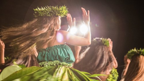 A group of dancers in traditional attire, wearing floral crowns, performing with vibrant movements against a dark background.
