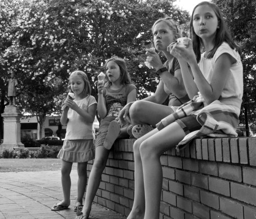 Four girls sitting on a brick wall, enjoying ice cream, with trees in the background. Black and white photo.