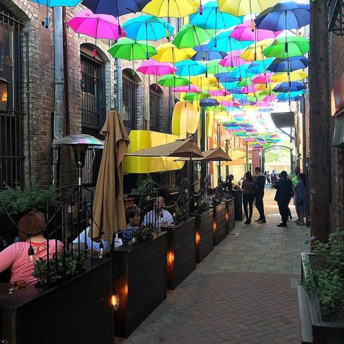 Colorful umbrellas hang overhead in a lively alleyway with outdoor dining and people enjoying the atmosphere.