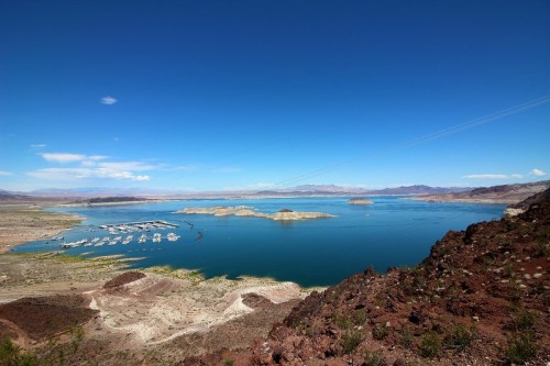 A panoramic view of a blue lake surrounded by rocky shores and distant mountains under a clear blue sky.