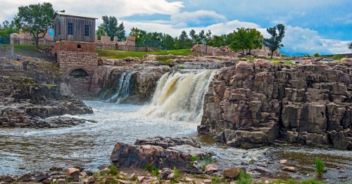A serene waterfall cascading over rocks, surrounded by trees and a distant building under a cloudy sky.