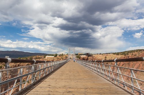 A wooden suspension bridge stretches across a canyon under a cloudy sky, with mountains in the background.