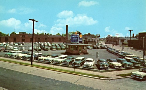 A vintage car dealership with numerous classic cars parked outside, featuring a large sign and a clear blue sky.