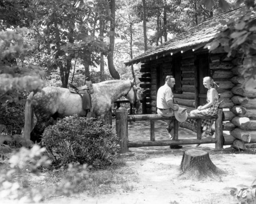 A man and woman sit on a fence near a log cabin, with a horse tied nearby, surrounded by trees.