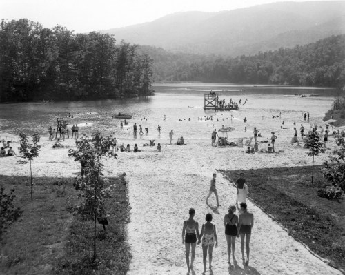 A black and white photo of a beach scene with people swimming, walking, and relaxing by a lake surrounded by trees.