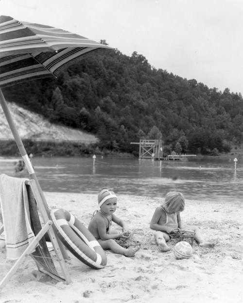 Two children playing on a sandy beach near a lake, with an umbrella and a diving platform in the background.