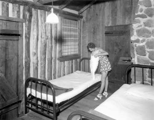 A woman adjusts a pillow on a bed in a rustic room with wooden walls and a stone accent.