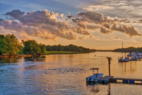 A serene river scene at sunset, with boats docked and colorful clouds reflecting on the water.