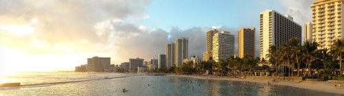 A panoramic view of a beach with high-rise buildings, palm trees, and people enjoying the water at sunset.