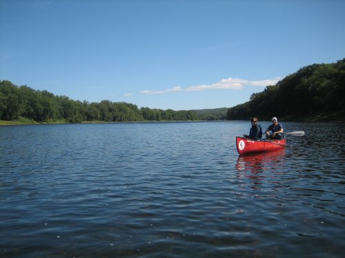 Two people in a red canoe paddling on a calm river surrounded by lush green trees under a clear blue sky.