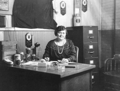 A woman sits at a desk in an office, writing and surrounded by documents and vintage items from the early 20th century.