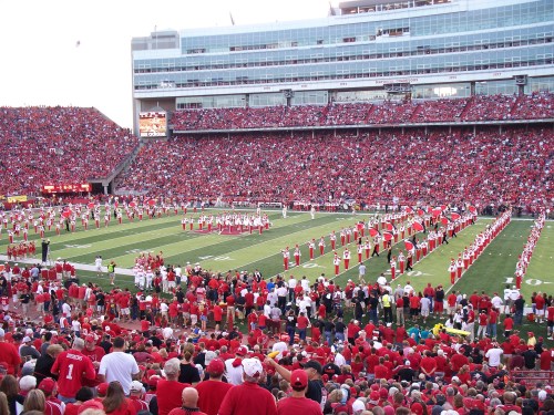 A packed stadium filled with fans in red, with a marching band performing on the field.