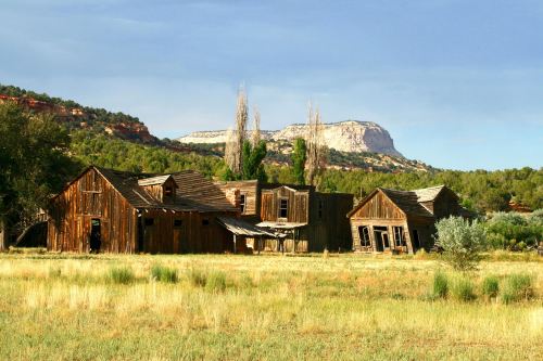 Abandoned wooden buildings in a grassy field, surrounded by trees and mountains under a clear blue sky.