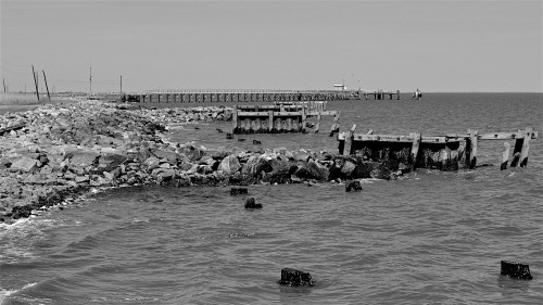 Black and white image of a rocky shoreline with a pier extending into the calm water under a clear sky.