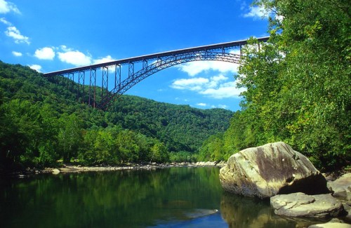 A scenic view of a large bridge spanning a river, surrounded by lush green hills and a clear blue sky.