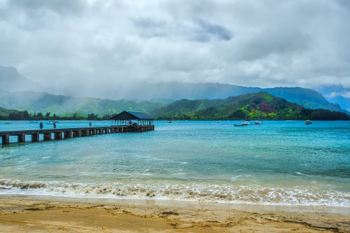 A serene beach scene with a wooden pier extending into turquoise waters, surrounded by lush green mountains under cloudy skies.