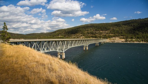 A long bridge spans a blue lake, surrounded by green hills and a partly cloudy sky. Golden grass lines the shore.