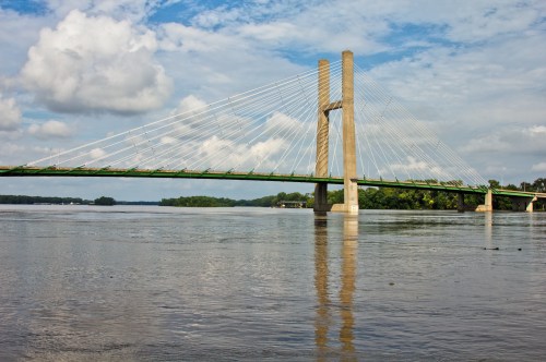 A cable-stayed bridge spans a river, reflecting in the water under a partly cloudy sky.