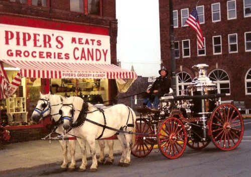 A horse-drawn fire wagon parked in front of a vintage grocery store with a sign reading "Piper's Meats Groceries Candy."