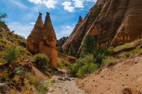Two unique rock formations rise amidst a desert landscape, surrounded by shrubs and under a blue sky with scattered clouds.