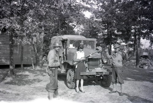 Two soldiers and a woman pose with a truck in a wooded area, one soldier holding a rifle.