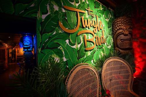 A vibrant wall with tropical leaves and the text "The Jungle Bird," featuring tiki decor and cozy seating.