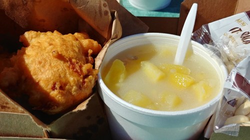 A bowl of creamy soup with chunks of pineapple next to a golden, fried fritter in a takeout container.