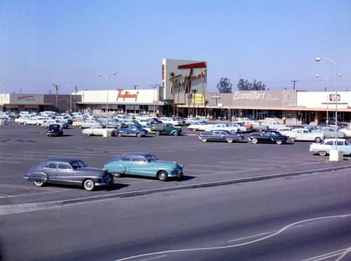 A vintage shopping plaza with a large parking lot filled with classic cars under a clear blue sky.