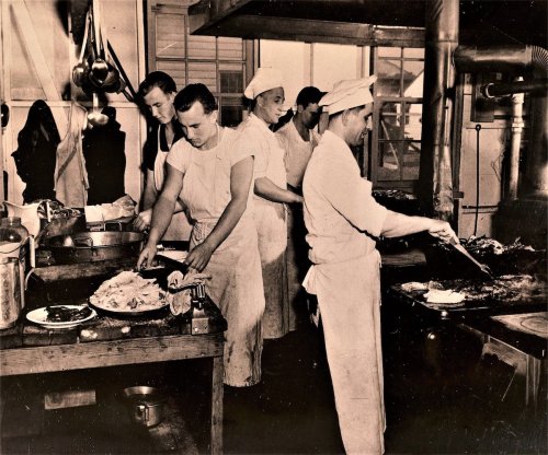 A vintage kitchen scene with chefs preparing food at large workstations, focused on their tasks.