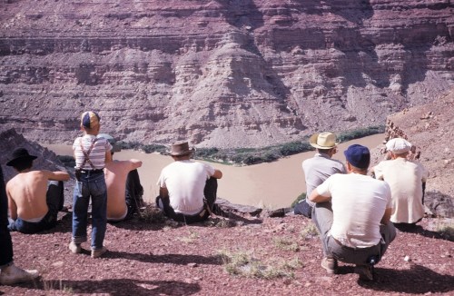 A group of people sitting on a rocky ledge, overlooking a river winding through a canyon.