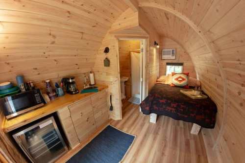 Cozy wooden interior of a small cabin featuring a bed, kitchenette, and bathroom area. Warm, inviting atmosphere.