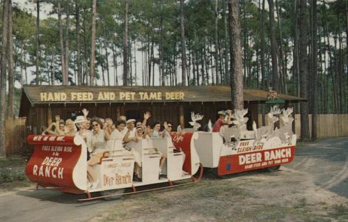 A vintage photo of a sleigh ride at a deer ranch, with people waving and surrounded by trees.