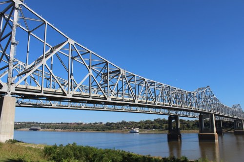 A steel truss bridge spans a river under a clear blue sky, with greenery along the water's edge.