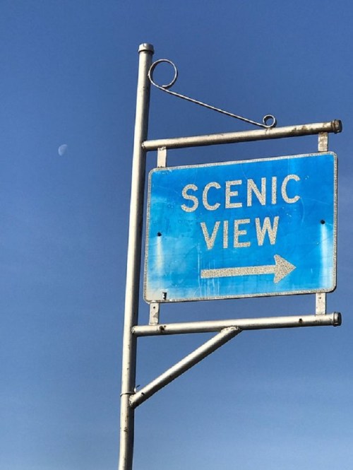 A weathered blue sign pointing right, reading "SCENIC VIEW," against a clear sky with a small crescent moon visible.