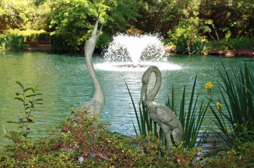 Two elegant heron statues by a serene pond with a fountain, surrounded by lush greenery and vibrant plants.