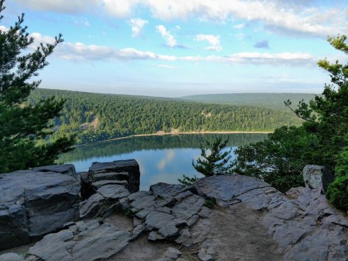 A scenic view of a calm lake surrounded by lush green forests and rocky terrain under a blue sky with clouds.