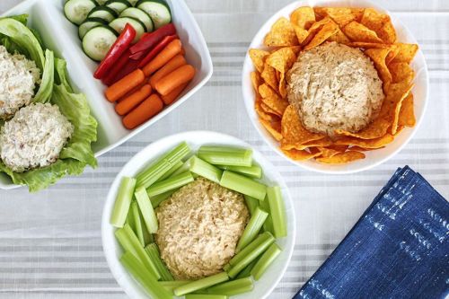 A table with three bowls of dip surrounded by fresh vegetables and chips, including carrots, cucumbers, and red peppers.
