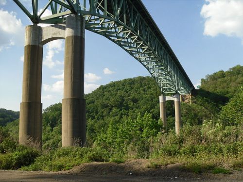 A large green bridge arches over a hillside, supported by tall concrete pillars, under a clear blue sky.
