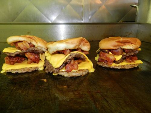 Three stacked cheeseburgers with bacon on a grill, featuring melted cheese and toasted buns.