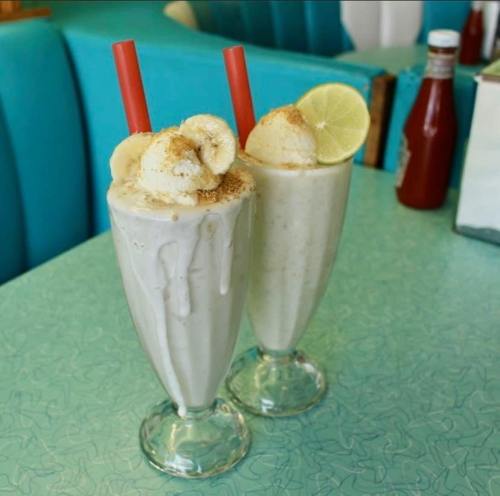 Two milkshakes in tall glasses, topped with banana slices and lime, served with red straws on a retro diner table.
